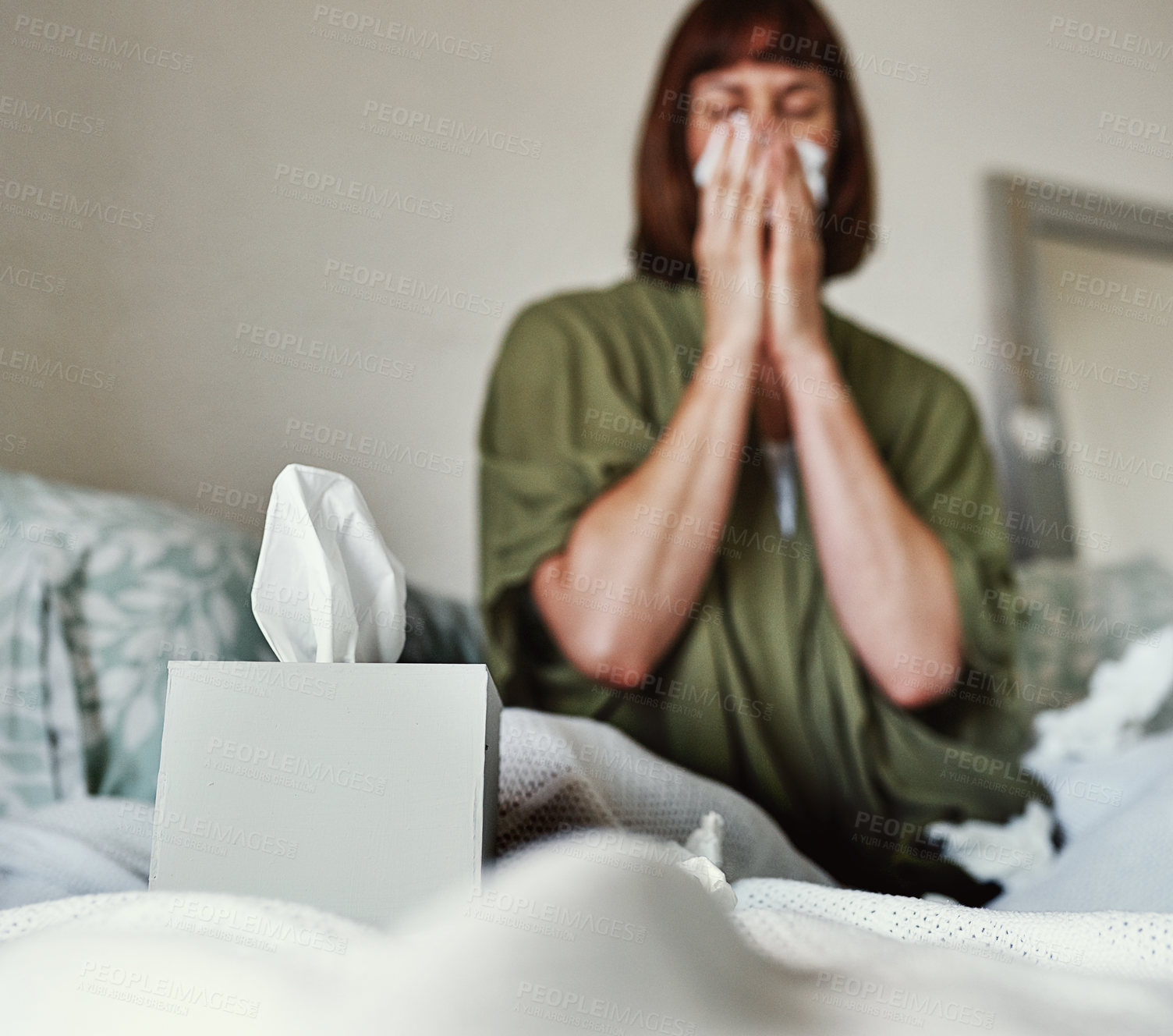 Buy stock photo Shot of a box of tissues placed on a bed with a woman blowing her nose in the background