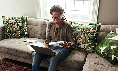 Buy stock photo Shot of a focused young man working on a laptop and doing paperwork while being seated on a sofa at home during the day
