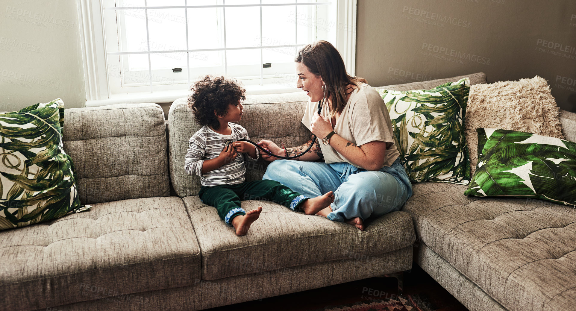 Buy stock photo Shot of a cheerful young woman and her son playing around with a stethoscope while being seated on a sofa at home during the day