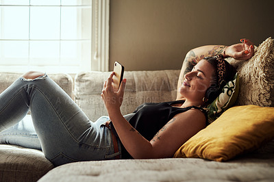 Buy stock photo Shot of a young woman using a smartphone and headphones on the sofa at home
