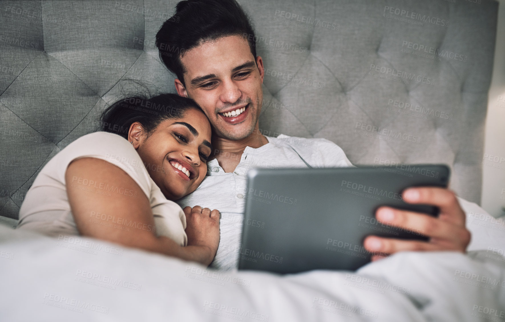 Buy stock photo Shot of a young couple looking at something on a digital tablet while lying in bed