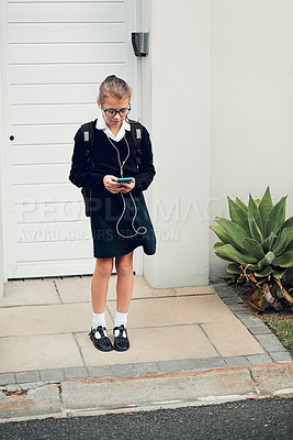 Buy stock photo Full length shot of a young schoolgirl listening to music and using her cellphone while standing outside her house