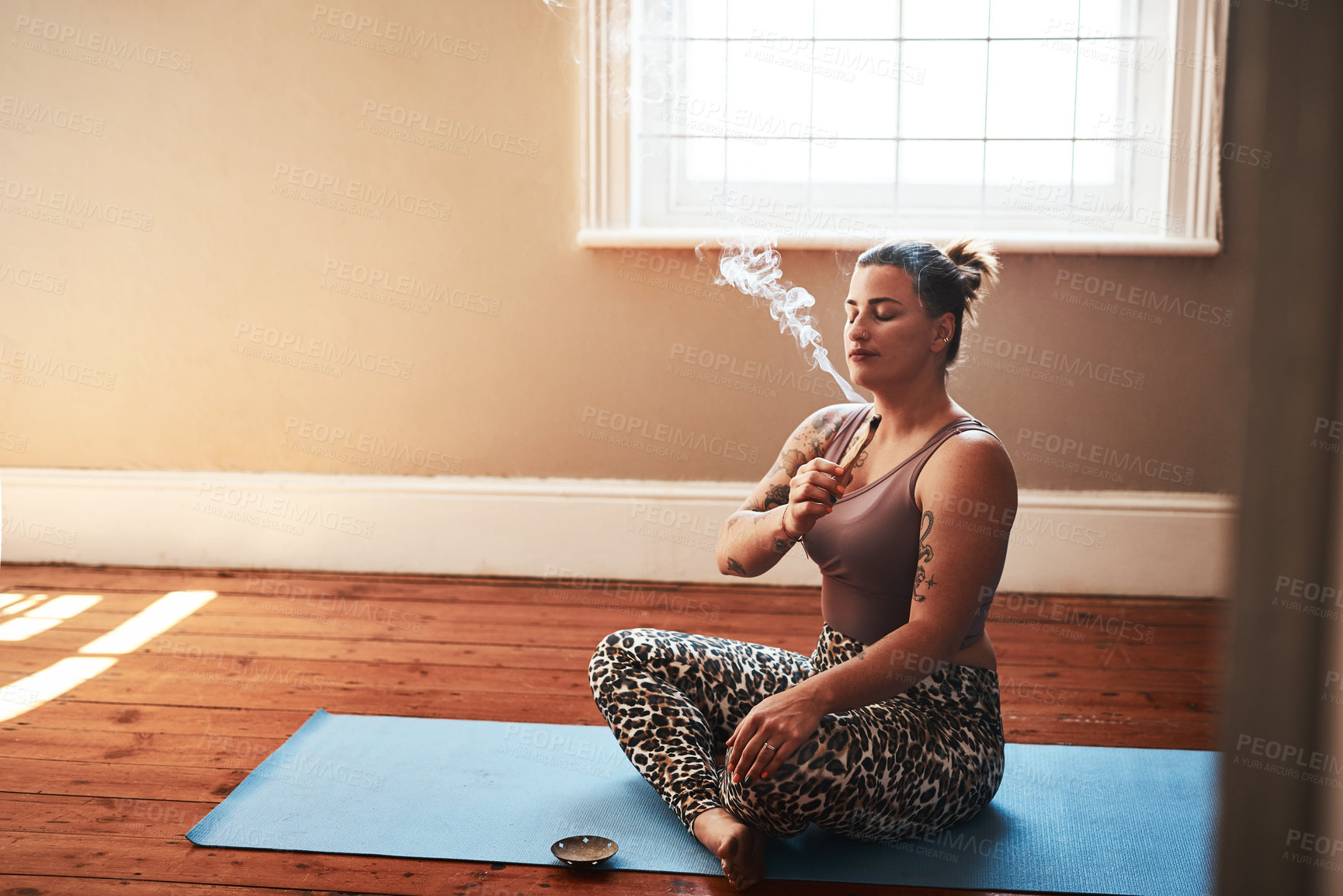 Buy stock photo Shot of a young woman burning a palo santo stick while sitting on a yoga mat at home