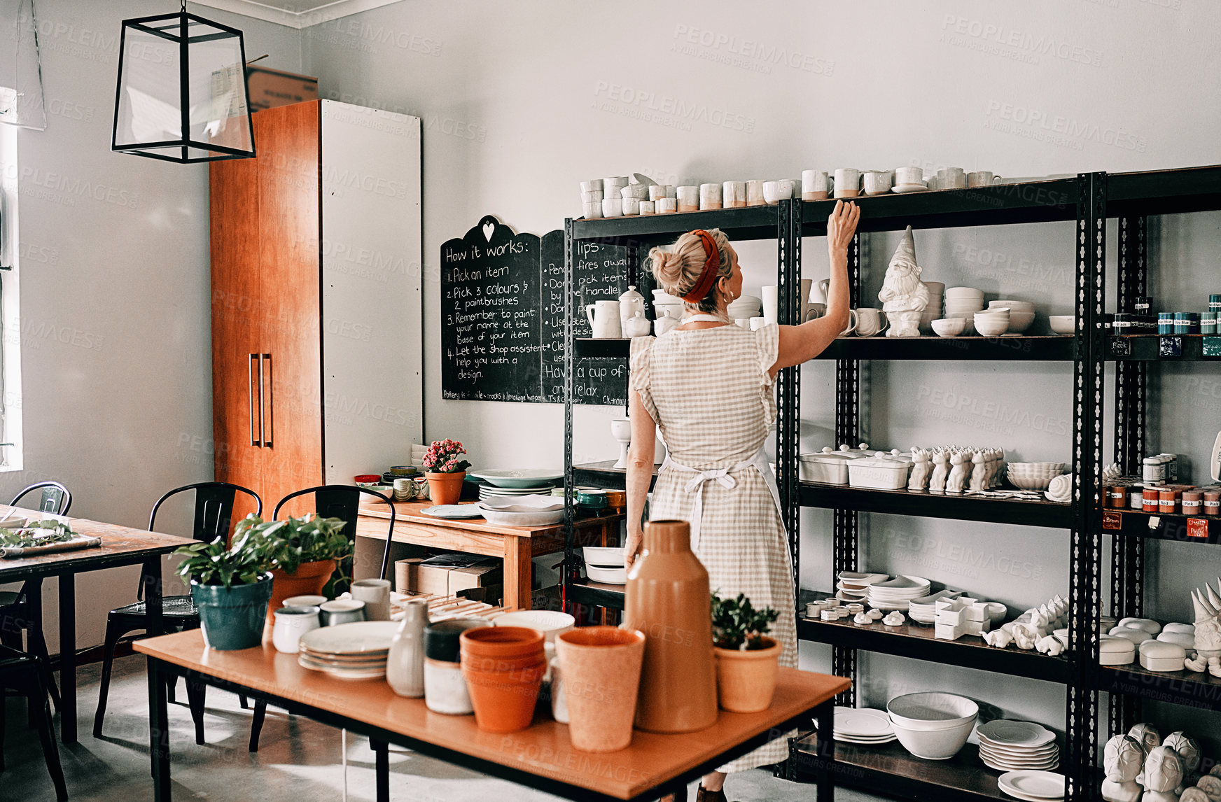 Buy stock photo Cropped shot of an unrecognizable woman standing and organising her pottery on a shelf in her workshop