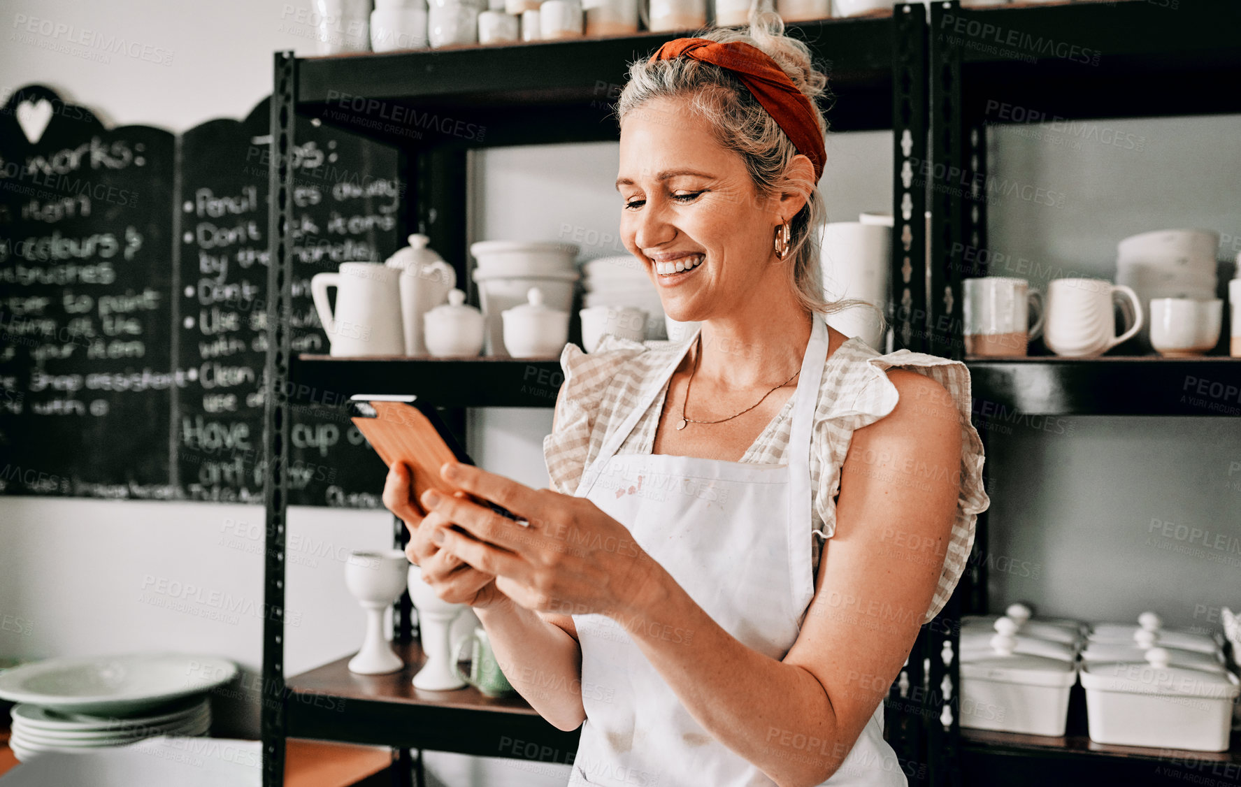 Buy stock photo Cropped shot of an attractive mature woman standing alone and using her cellphone in her pottery workshop