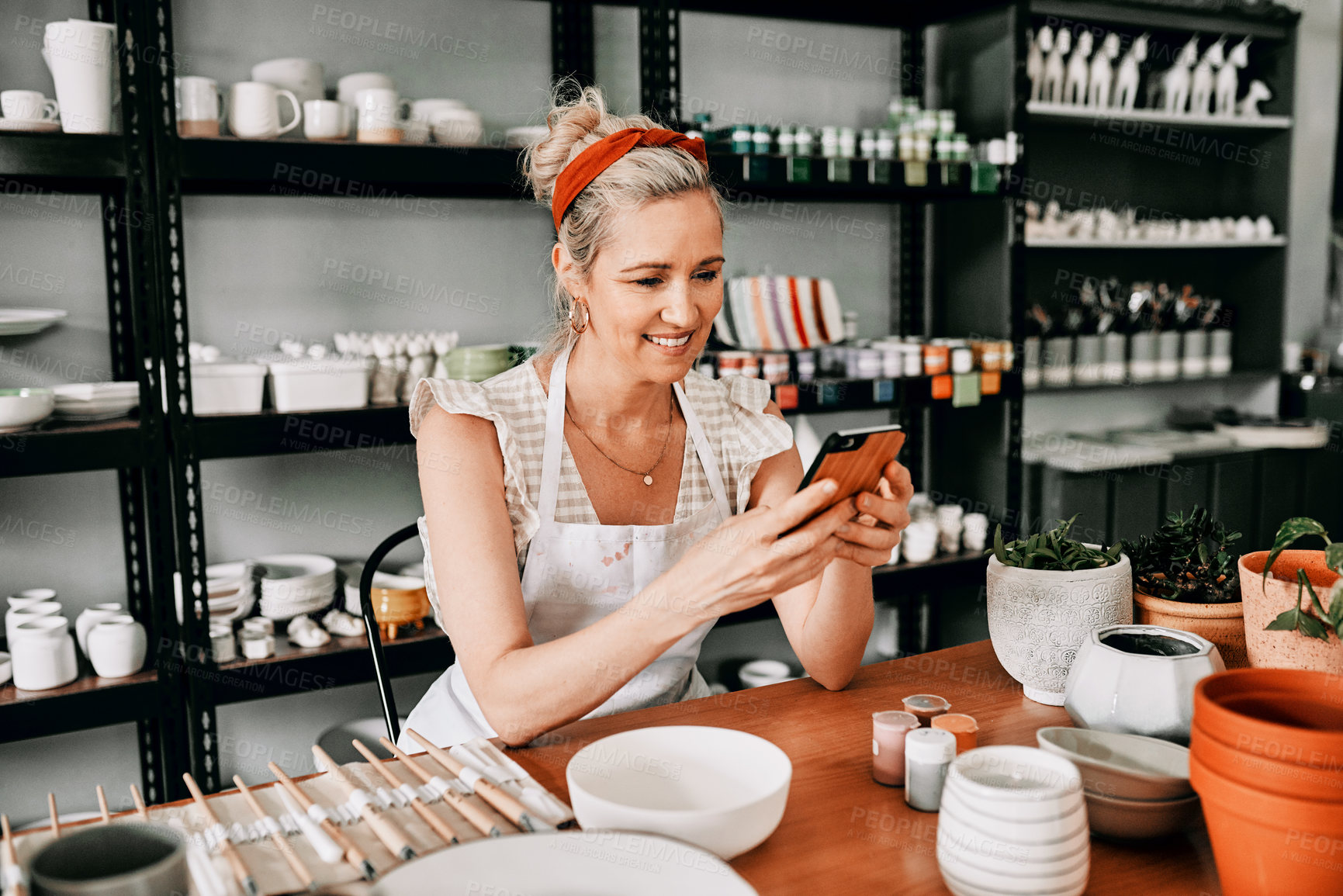 Buy stock photo Cropped shot of an attractive mature woman sitting alone and using her cellphone in her pottery workshop