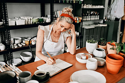 Buy stock photo Cropped shot of an attractive mature woman sitting alone in her pottery workshop and sketching in a notebook