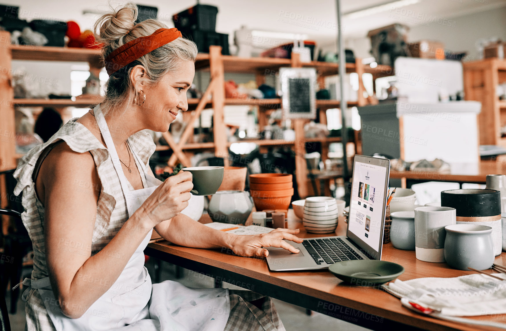 Buy stock photo Cropped shot of an attractive mature woman sitting alone and using her laptop in her pottery workshop while drinking coffee