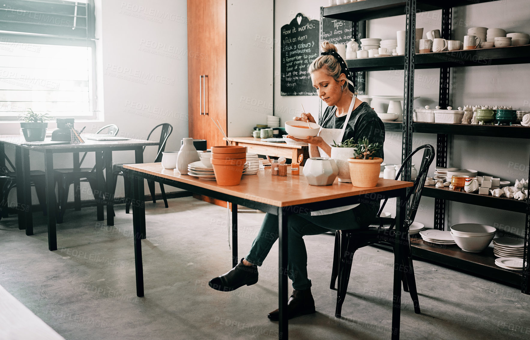 Buy stock photo Full length shot of an attractive mature woman sitting alone and painting a pottery bowl in her studio