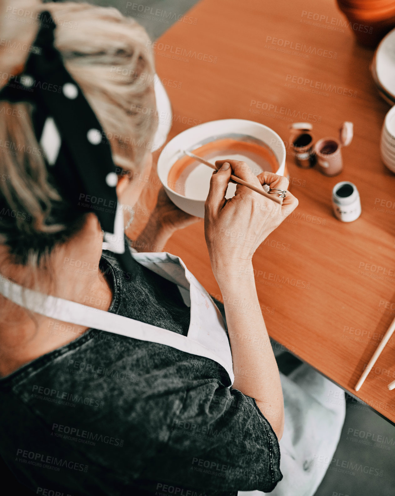 Buy stock photo High angle shot of an unrecognizable woman sitting alone and painting a pottery bowl in her workshop