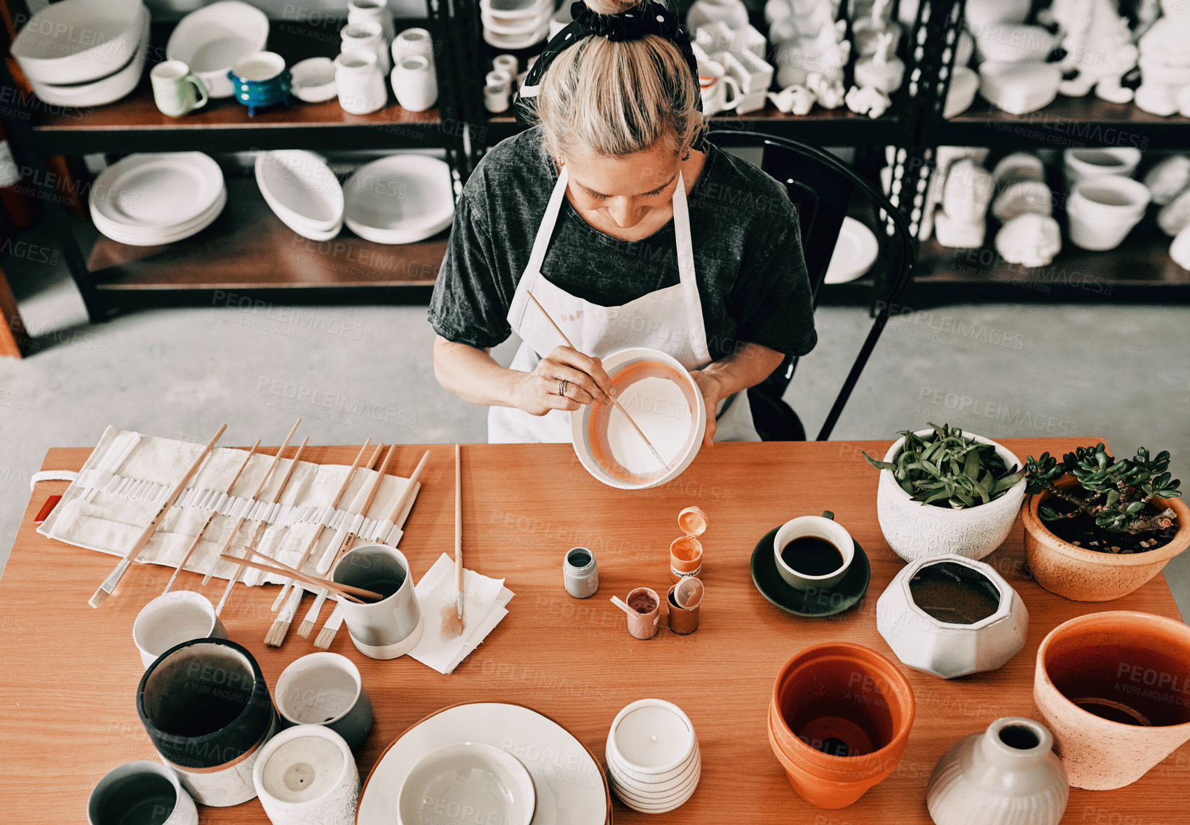 Buy stock photo High angle shot of an unrecognizable woman sitting alone and painting a pottery bowl in her workshop