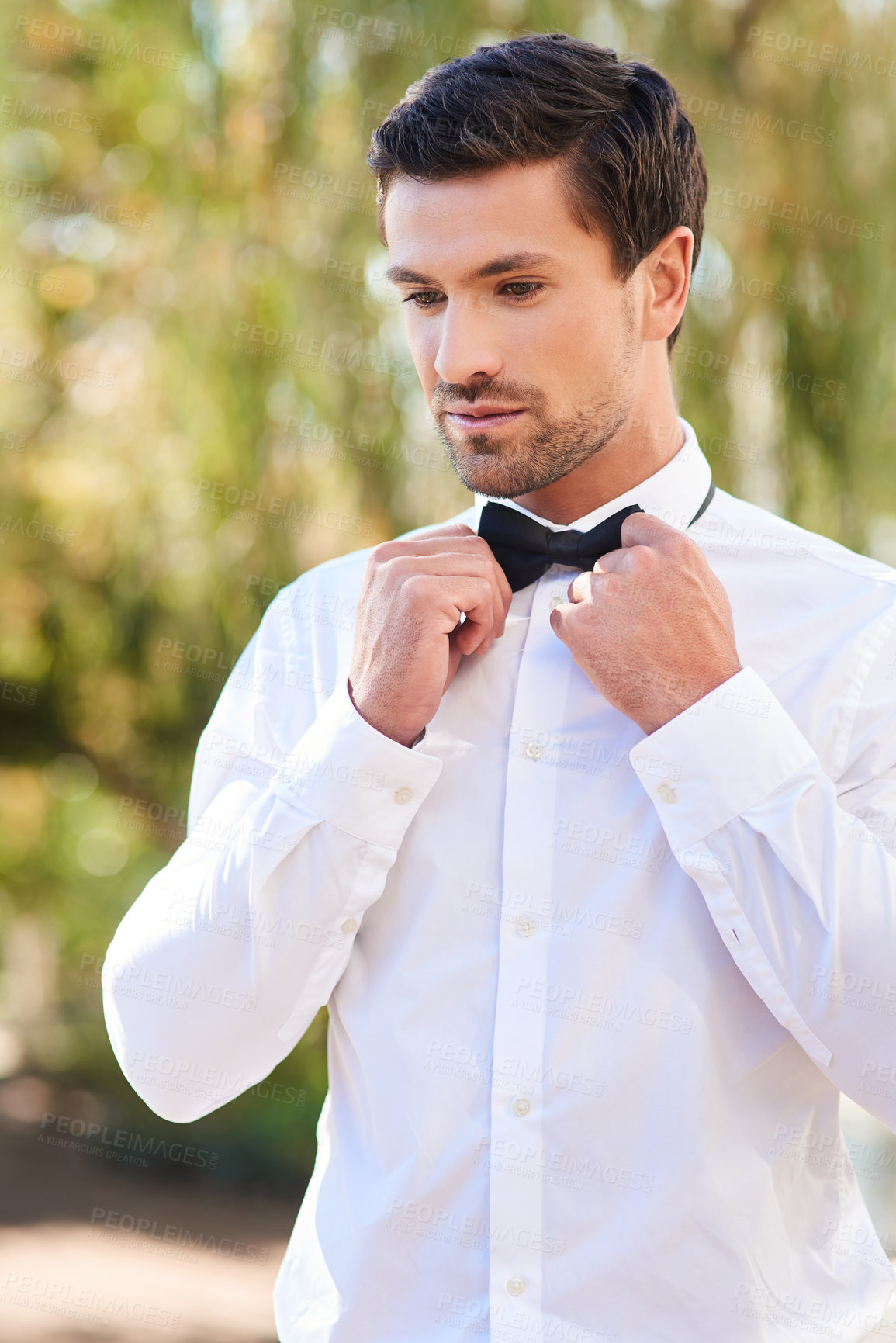 Buy stock photo Shot of a handsome young bridegroom putting on a bowtie while standing outdoors on his wedding day