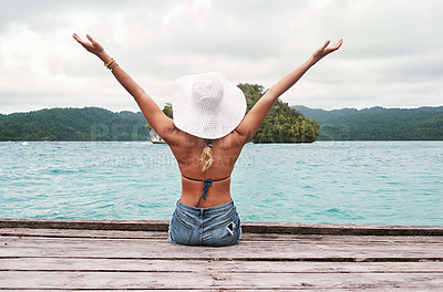 Buy stock photo Rearview shot of an unrecognizable young woman seated on a jetty while lifting her hands in the air in excitement outside next to the ocean during the day
