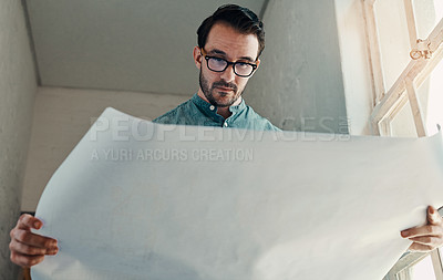 Buy stock photo Cropped shot of a handsome young male architect looking at a blueprint while standing in an office