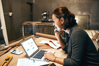 Buy stock photo Cropped shot of an attractive young female carpenter working on her laptop while talking on her cellphone inside of a workshop