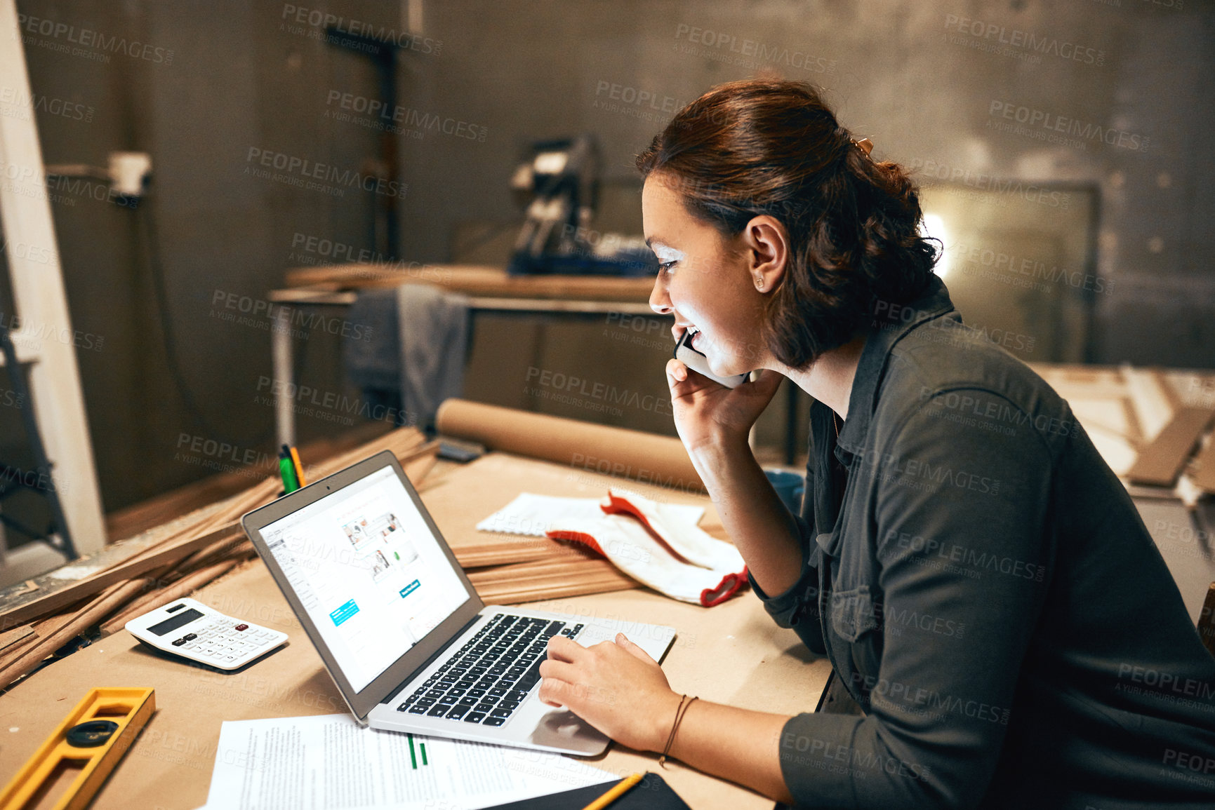 Buy stock photo Cropped shot of an attractive young female carpenter working on her laptop while talking on her cellphone inside of a workshop