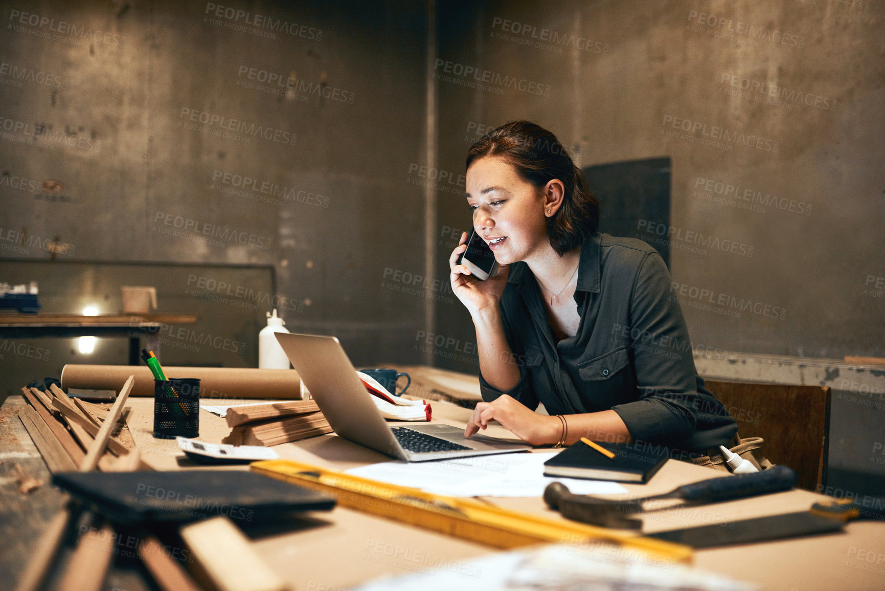 Buy stock photo Cropped shot of an attractive young female carpenter working on her laptop while talking on her cellphone inside of a workshop