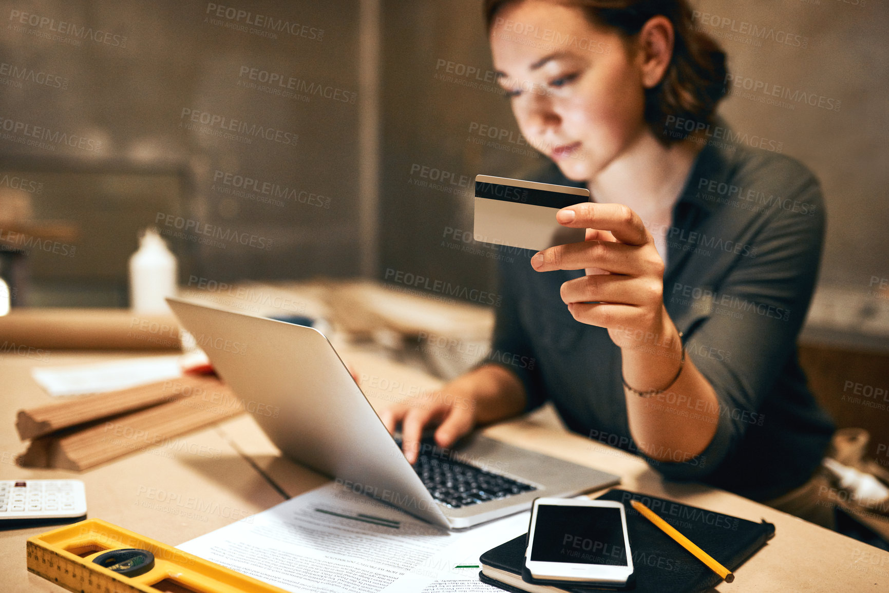 Buy stock photo Cropped shot of an attractive young female carpenter holding her credit card while doing online shopping inside of a workshop at night