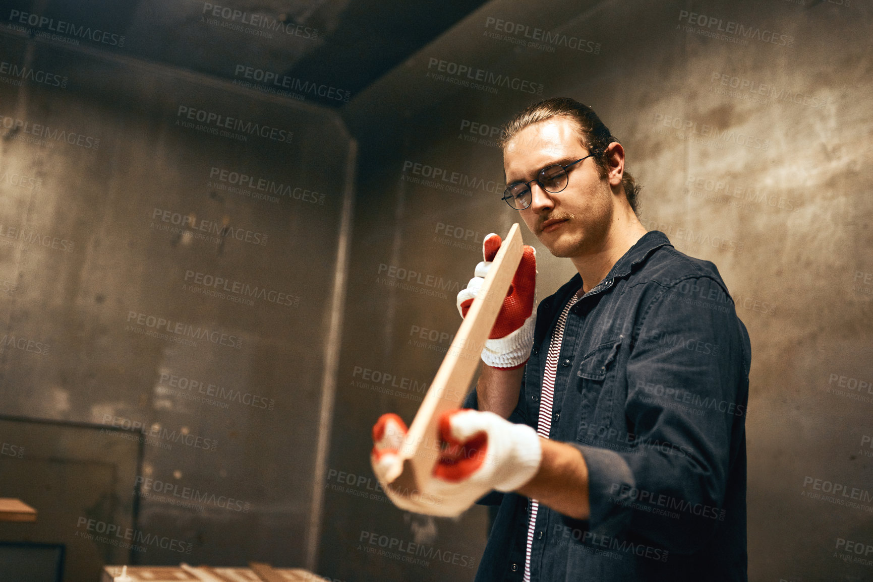 Buy stock photo Cropped shot of a focused young male carpenter holding and examining a piece of wood while working inside of a workshop at night