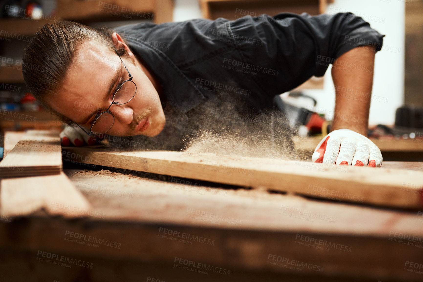 Buy stock photo Cropped shot of a focused young male carpenter blowing dust off of a piece of wood after sanding it inside of his workshop during the night