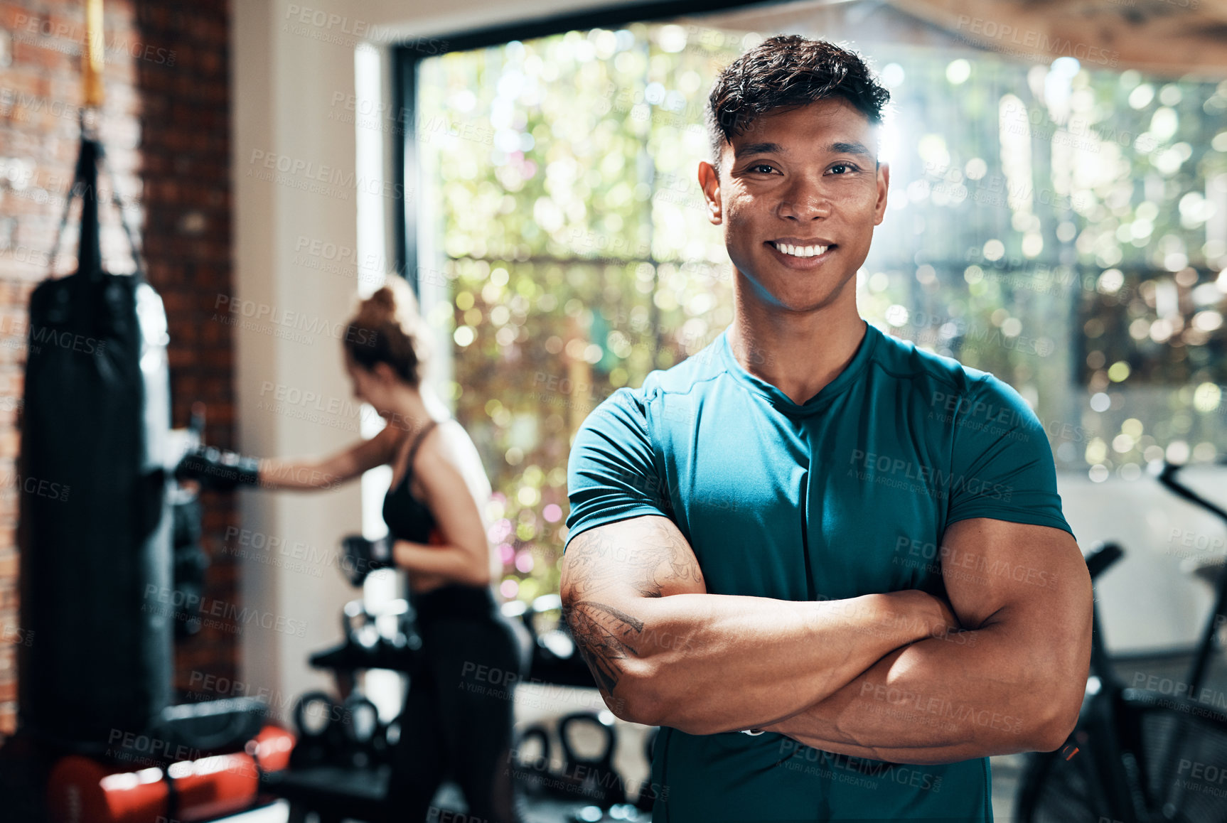 Buy stock photo Cropped shot of a handsome young sportsman standing with his arms crossed in a gym with his fitness partner in the background