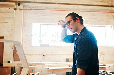 Buy stock photo Shot of a handsome young carpenter wiping sweat off his forehead while working hard inside a workshop
