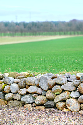 Buy stock photo Closeup of a stone wall built with rocks along an open green field on a farm. A rocky barrier between a dirt road and a meadow or pasture. Manmade structure with natural landscape in the background