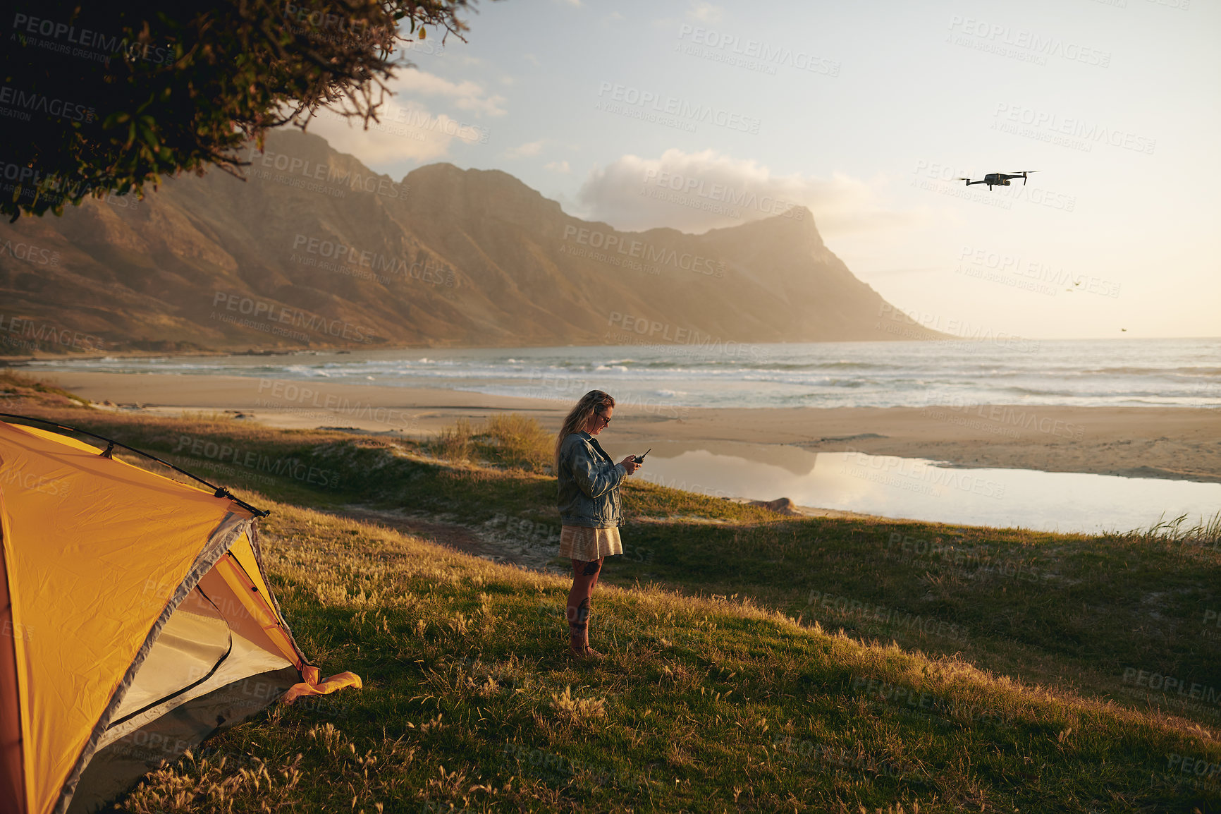 Buy stock photo Full length shot of an attractive young woman flying a drone while camping outdoors by herself near the beach