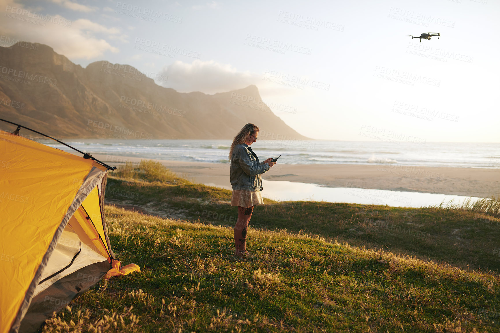Buy stock photo Full length shot of an attractive young woman flying a drone while camping  outdoors by herself near the beach