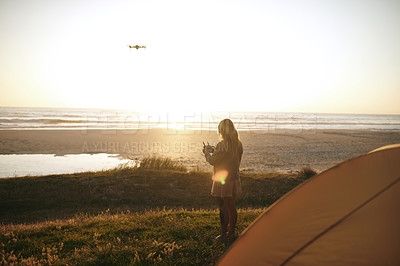 Buy stock photo Full length shot of an unrecognizable young woman flying a drone while camping outdoors by herself near the beach