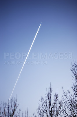 Buy stock photo Jet airplane contrail above leafless trees against a clear blue sky background with copyspace. View of a distant passenger plane flying at high altitude leaving a long white smoke trail behind