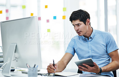 Buy stock photo Shot of a young businessman writing notes while using a digital tablet and computer in an office
