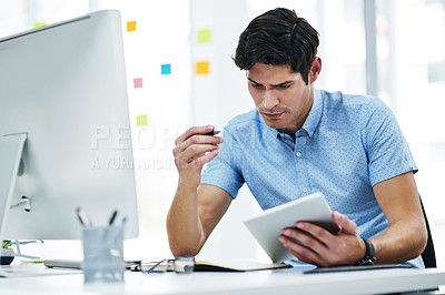 Buy stock photo Shot of a young businessman writing notes while using a digital tablet and computer in an office