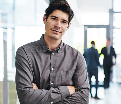 Buy stock photo Cropped portrait of a handsome young businessman standing with his arms folded in an office with his colleagues in the background
