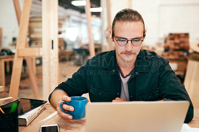 Buy stock photo Cropped shot of a handsome young carpenter drinking coffee and working on a laptop inside his workshop