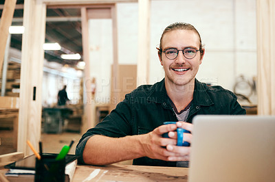 Buy stock photo Portrait of a handsome young carpenter drinking coffee and working on a laptop inside his workshop