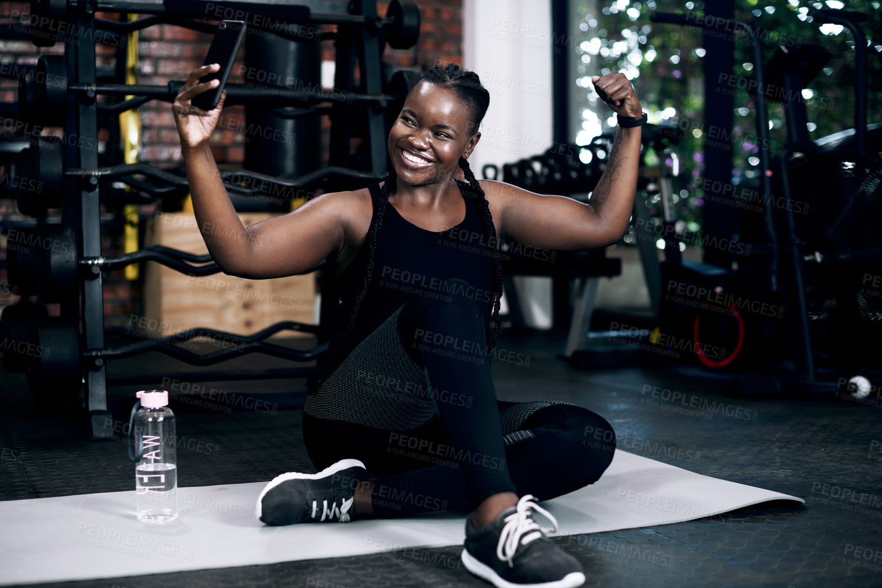 Buy stock photo Full length shot of an attractive young female athlete flexing her biceps while sitting on her towel in the gym