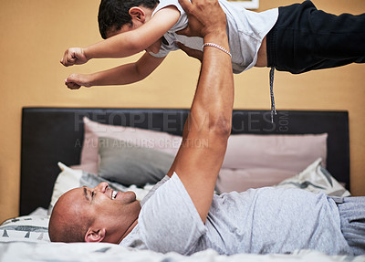 Buy stock photo Cropped shot of a cheerful young man lifting up his son in the air to fly like a super hero while lying down on a bed at home during the day