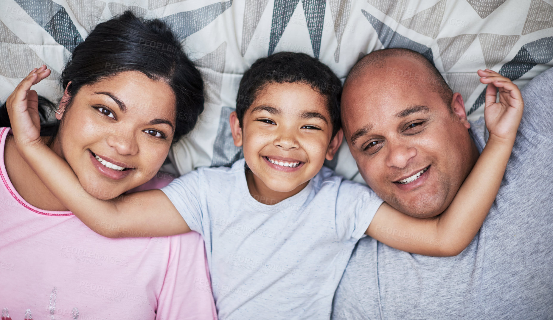 Buy stock photo Portrait of a cheerful young family lying down and hanging out in bed smiling all the way at home during the day