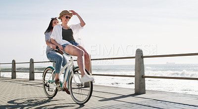 Buy stock photo Full length shot of a happy young couple riding a bicycle together on a promenade near the beach