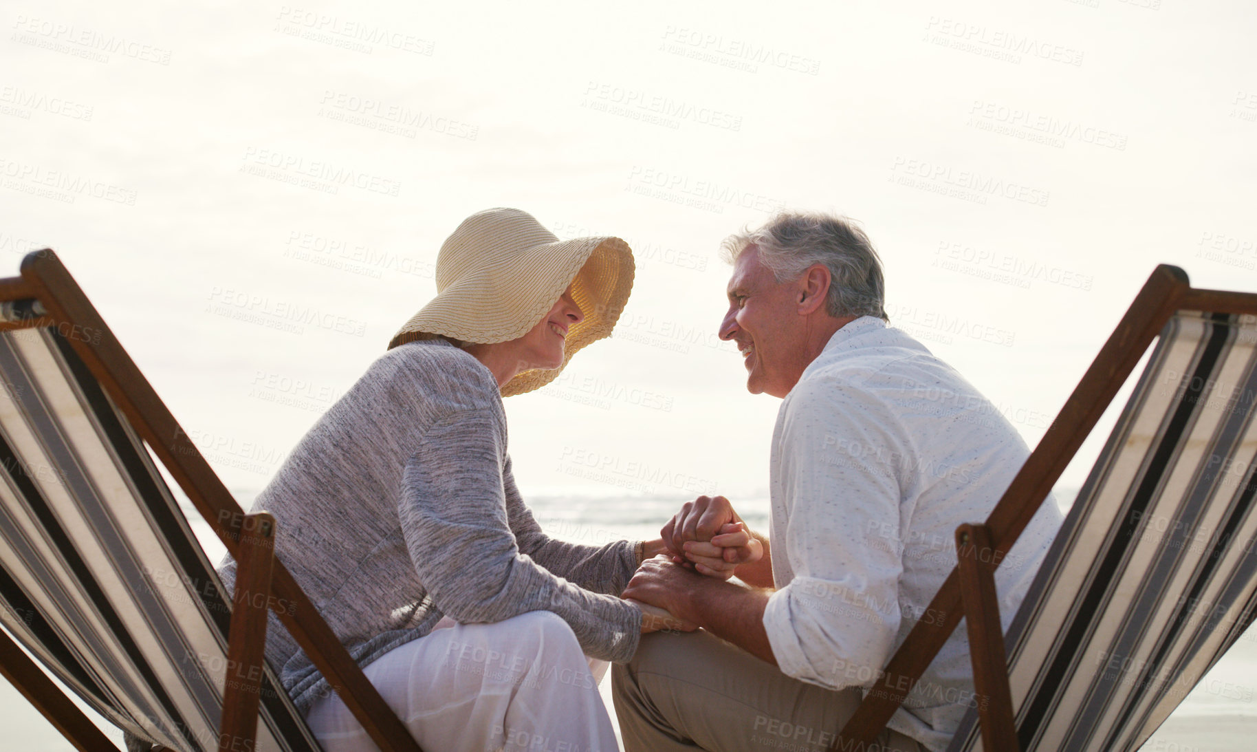 Buy stock photo Cropped shot of an affectionate senior couple holding hands intimately while sitting on loungers at the beach