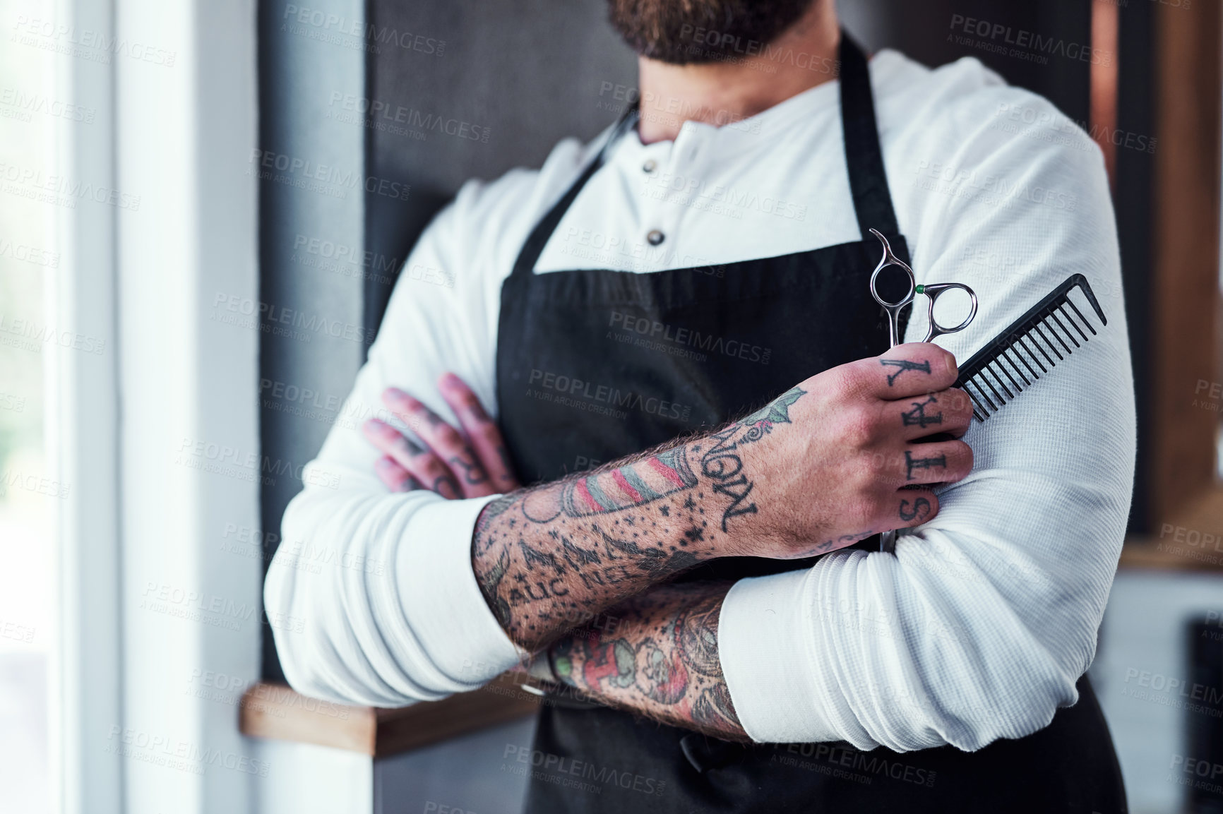 Buy stock photo Cropped shot of a unrecognizable tattooed barber posing with a pair of scissors and a hair comb inside a barbershop