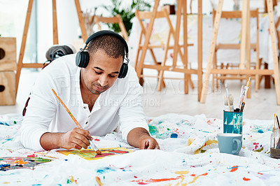 Buy stock photo Cropped shot of a handsome young artist sitting alone and painting while listening to music through headphones