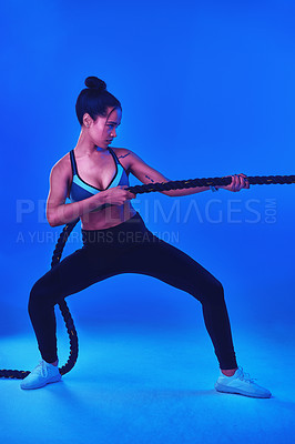 Buy stock photo Full length shot of an attractive young sportswoman working out with battle ropes against a blue background