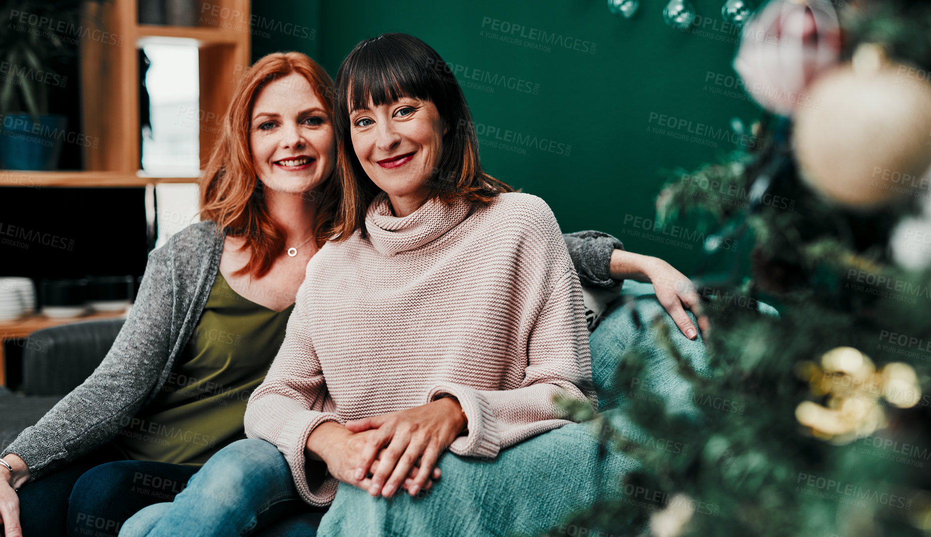 Buy stock photo Portrait of two attractive middle aged women sitting together on a sofa next to a Christmas tree at home