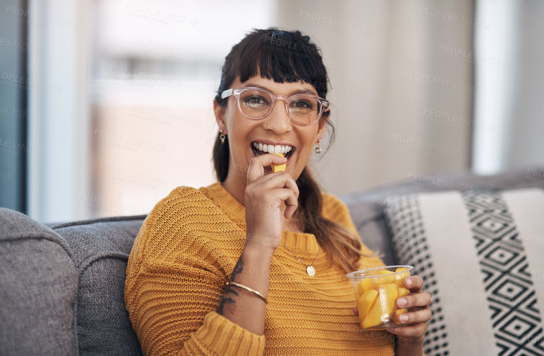 Buy stock photo Cropped portrait of an attractive young woman sitting alone in her living room and enjoying mangoes during a day off