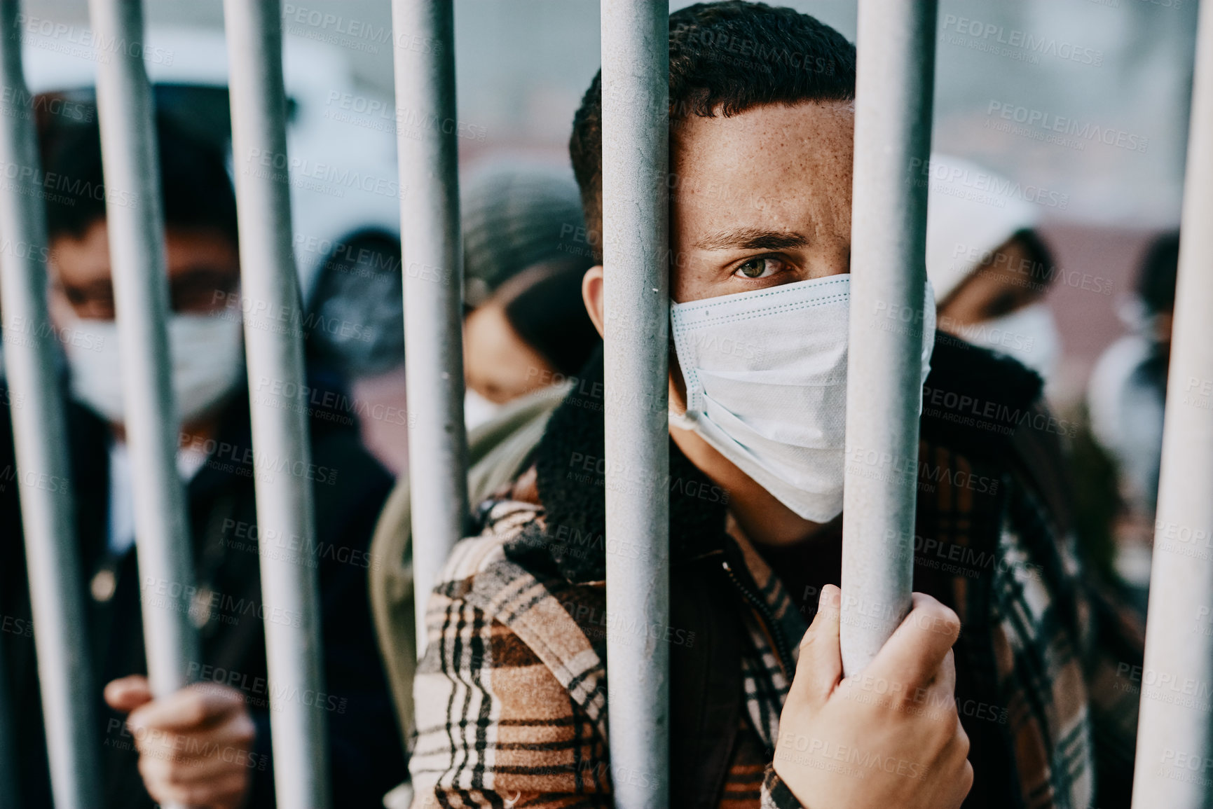 Buy stock photo Shot of a young man wearing a mask while stuck behind a gate in a foreign city