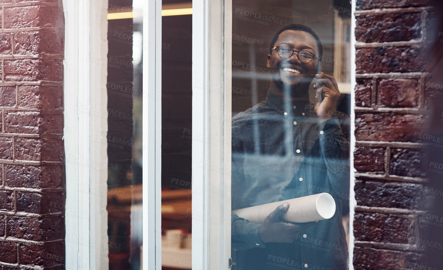 Buy stock photo Man, architect and phone call in office with documents in roll, blueprint and negotiation with contractor. African person, smartphone and smile by window in conversation, floor plan and decision