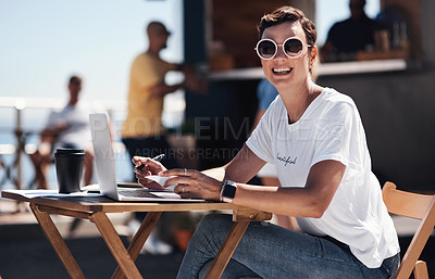Buy stock photo Portrait of a cheerful young woman working on her laptop while being seated at a table next to a beach promenade outside during the day