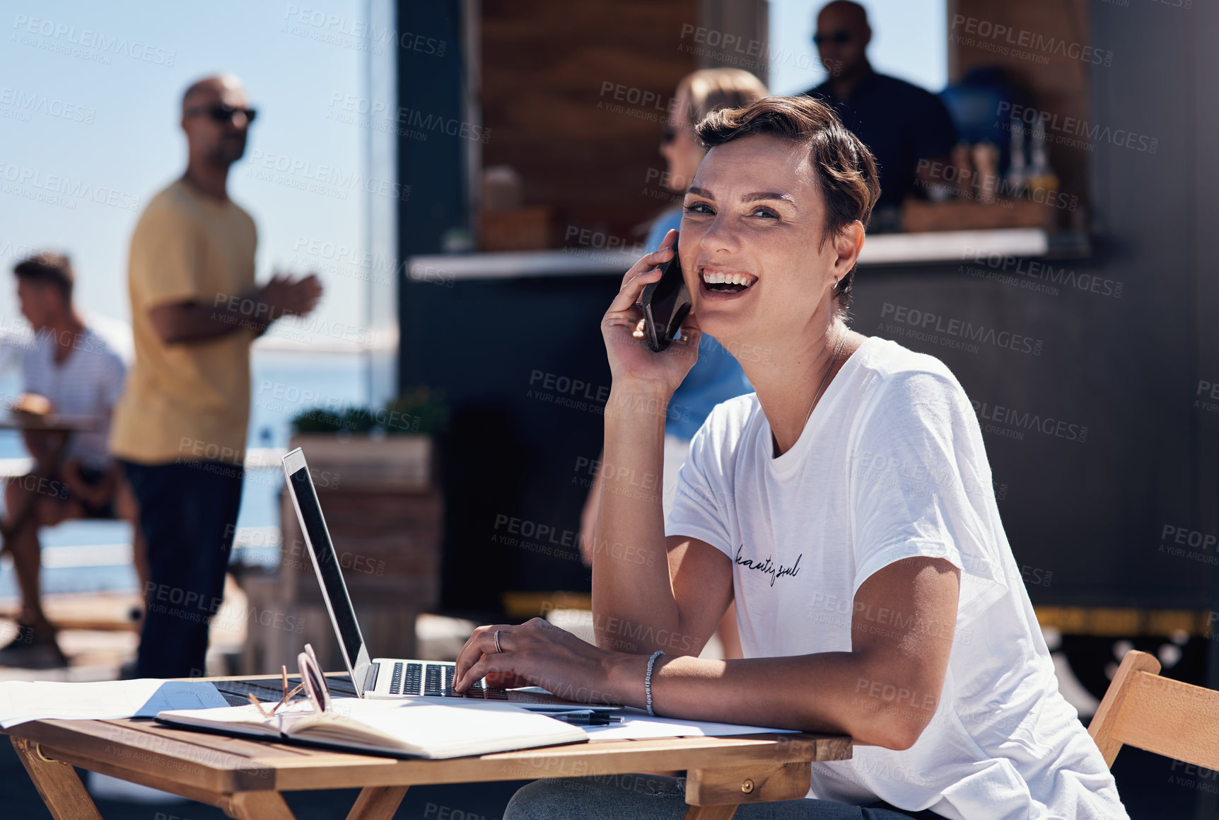 Buy stock photo Portrait of a cheerful young woman talking on her cellphone while doing work on her laptop next to a beach promenade outside during the day